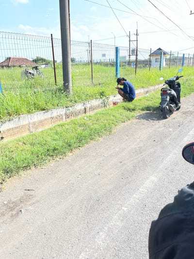 A young man with dark hair and a black cap, wearing a blue long-sleeved shirt and dark shorts, is crouching on the grassy shoulder of a dirt road. He is looking at his phone, which he holds in his hands. Next to him, a red and white bucket is on the ground. A dark-colored motorcycle is parked on the road to his right. In the background, a chain-link fence runs along the edge of a field of tall green grass. Beyond the fence, there is a building with a yellow roof and a smaller white structure, as well as a house with a red tiled roof. A large concrete utility pole stands on the left side of the frame. The sky is clear and blue with some scattered clouds. The scene appears to be located in a rural or semi-rural area in Pamotan, Indonesia, given the context. The lighting suggests it is daytime, likely late morning or afternoon, due to the strong shadows.