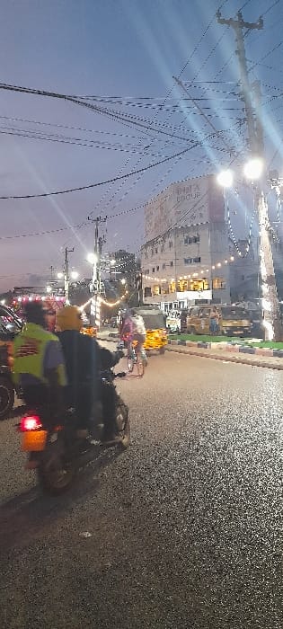 The image depicts an urban street scene in Majengo ward, Kenya, at dusk or night. The asphalt road in the foreground appears wet or reflective under artificial illumination. A motorcycle, positioned in the lower-left quarter of the frame, travels away from the viewer. It carries two individuals: the driver wears a yellow helmet, and the passenger wears a yellow-green reflective vest. The motorcycle's red tail light is illuminated. Further down the road, a person on a bicycle moves in the same direction. On the right side of the road, multiple yellow tuk-tuks (auto-rickshaws) are visible, some parked or idling. The street is lined with utility poles supporting a dense network of overhead wires. Several bright streetlights are active, casting significant glare. In the midground, a multi-story building displays illuminated windows and a partially visible sign that reads "MHA GROUP" on its upper facade. Decorative string lights are strung between structures along the street. The sky above is a deep twilight blue-grey.