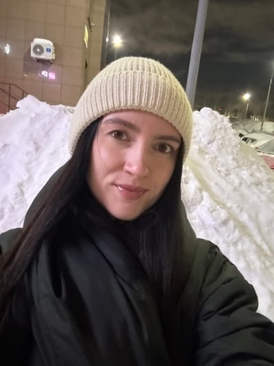 A woman wearing a light beige knit hat and a dark puffer jacket poses outdoors at night. She has dark hair and a fair complexion. Piles of snow are visible in the background, and there are buildings, a street lamp, and what appears to be parked cars. An air conditioning unit is mounted on the wall behind her. The sky is dark, suggesting it's late at night or very early morning.