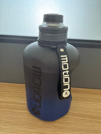 A black and blue gradient water bottle with the word "motion" printed vertically on its side in white, embossed lettering is shown in a close-up, eye-level, studio shot. The bottle has a black lid and a black carrying strap attached to its neck, also featuring the "motion" logo. The bottle is placed on a light brown wooden surface, possibly a table, in front of a gray and white textured wall. The background is blurred, with a faint light source visible. The scene does not depict any specific location in Umm Salal Ali, Qatar, or any people.