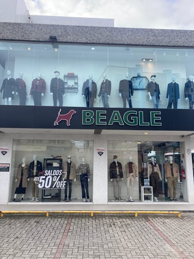 This image captures the exterior of a clothing store named "BEAGLE" on an overcast day in Florianópolis, Brazil. The store occupies the ground floor of a modern building with a distinct tiled facade. The main feature is a large, glass display window showcasing numerous male mannequins dressed in various fashion items, including suits, jackets, and casual wear.
The store's name, "BEAGLE," is vertically displayed in large green letters on a dark panel separating two sections of the glass display, accompanied by a red beagle dog logo. The left section of the window features mannequins alongside a large, decorative wall clock. The right section displays more mannequins, some wearing patterned shirts and others in neutral-toned outerwear. A prominent white sign in this window announces "SALDOS ATÉ 50% OFF" (Sales up to 50% Off), with smaller text indicating the promotion is "VÁLIDA PARA COLEÇÕES 064 E ANTERIORES" (Valid for collections 064 and previous).
At ground level, glass doors lead into the store, with reflections visible. A "Khronos 24h" security sticker is present near the entrance, along with partial text that appears to be related to store information, possibly "FUNCIONAMENTO" (Operating Hours). The paved sidewalk in front of the store features light-colored interlocking blocks, a red painted stripe, and a weathered yellow metal railing running parallel to the building. The sky is largely obscured by clouds, suggesting an overcast daytime. The visible Portuguese text serves as a strong location cue.