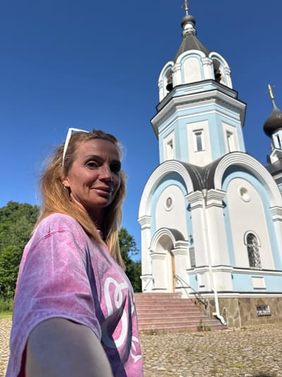 A blonde woman wearing a pink tie-dye t-shirt and white sunglasses on her head stands in front of a light blue and white church with a bell tower. The church has a black dome and a gold cross on top. The woman is smiling and looking at the camera. The ground in front of the church is paved with cobblestones. There are trees in the background. The sky is clear and blue. This is a selfie taken in front of the Church of the Assumption of the Blessed Virgin Mary in St. Ozerki, Russia.