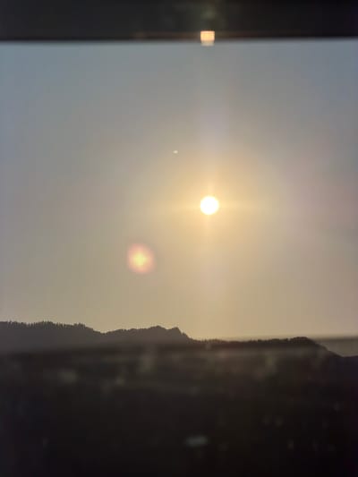 The image depicts a hazy, sunlit sky with the sun appearing as a bright orb, slightly diffused by atmospheric conditions. To the right, the silhouette of a treeline or land formation is visible against the bright sky. The overall scene suggests an outdoor environment, potentially a landscape. There are no people or discernible objects, and no text is present. The quality of light indicates it is likely either sunrise or sunset, or a heavily overcast day with the sun obscured. The presence of atmospheric haze contributes to a muted, soft visual. The location context of Elekmonar, Russia, does not provide specific visual cues within the image itself, but suggests a northern latitude environment.