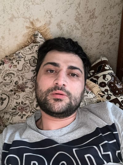 A man lies on a sofa, resting his head on a patterned cushion. The man has dark hair, a beard, and light-colored eyes. He is wearing a gray t-shirt with a large black and white graphic design on the right side. The sofa is covered in a textured fabric. Behind the man, there are two patterned cushions, one with tassels. The lighting suggests it is daytime. The scene appears to be indoors.