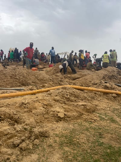 The image shows a large group of people congregated on a dirt embankment or hillside, likely in Maiyanga, Nigeria. The ground is bare earth, sparsely covered with grass in some areas. A yellow pipe runs vertically along the side of the embankment. The sky is overcast. Individuals are dressed in a variety of clothing, with some wearing work vests or carrying items on their heads. Some people are standing on top of each other.
