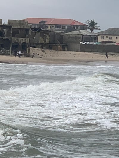The image captures a coastal landscape in Ibeju, Nigeria, under an overcast sky. The foreground is dominated by a grey ocean with multiple breaking waves exhibiting white foam. A sandy beach strip occupies the midground.
Along the immediate background of the beach, a series of structures are visible. On the left, several multi-story concrete buildings appear to be in various states of disrepair or incomplete construction, displaying exposed rebar and deteriorated facades. Two individuals are present on the beach near these structures; one stands, while another is seated or crouched on the sand.
To the center-right, a single-story concrete building with a flat, grey roof stands closer to the beach, with laundry items hanging on an adjacent wall. Further inland, a larger multi-story building with a distinctive red corrugated metal roof is prominent, flanked by a beige-colored multi-story building with a dark roof to its right. A solitary palm tree rises behind these buildings. A concrete wall runs along the rear of the sandy area.
On the far right of the beach, a single individual is observed walking along the water's edge, heading away from the primary cluster of buildings. No discernible interactions between people are evident. The scene portrays a blend of natural coastal elements and varied human habitation.