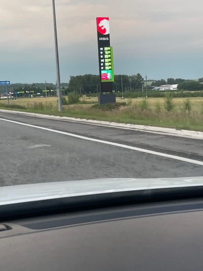 The image shows a digital gas price sign for "IRBIS" in Кощаково, Russia. The sign displays prices for various fuel types, including 92 octane gasoline at 49.00, 95 octane gasoline at 52.00, diesel at 53.90, and 100 octane gasoline at 54.90. The background features a gas station with multiple pumps, a road, and a cloudy sky. There are no people visible in the image.