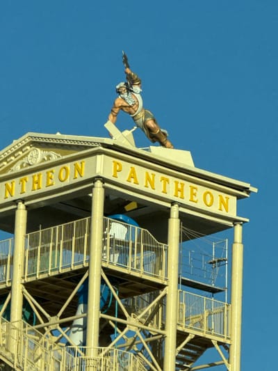 The image shows a close-up view of the upper section of a tower-like structure against a clear blue sky. At the very top of the structure is a statue of a muscular male figure, likely a god or hero from Greek mythology, poised as if in action, holding a weapon. The statue is painted in realistic tones of brown, white, and gold.

Below the statue is a pedimented roof-like structure with decorative carvings and the golden text "PANTHEON" prominently displayed. The architecture appears to be inspired by classical Greek design, with columns supporting the roof.

The rest of the image is dominated by the tower itself, which is constructed with a yellow metal framework. Multiple levels are visible, featuring walkways with yellow railings and what appear to be sections of a water slide with blue and white elements visible through the structure. Scaffolding is also present on one side of the tower. The overall impression is of an amusement park attraction or a themed building. The location is identified as Chersonisos Municipal Unit, Greece.