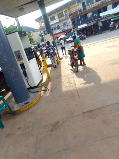 The scene is a gas station in Orlu, Nigeria. The foreground shows a concrete surface with visible cracks and imperfections. A red motorcycle is parked next to a woman in a blue and gold dress. She appears to be holding bottles of water. To the left, there are gas pumps with digital displays, set on a slightly raised concrete pad. A few individuals are standing near the pumps, including one man wearing a black and white jersey with the number "16" on the back. In the background, buildings line the street, one with a blue awning, and street lights are visible. Motor vehicles are present on the roadway.