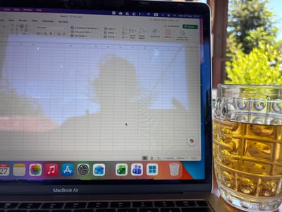 The image shows a MacBook Air open, displaying a spreadsheet program, likely Excel, with "Book2 - No Label" at the top. The date "Wed 27 May 13:31" is visible in the upper right corner of the screen. Below the screen are application icons in the dock, including Calendar, Photos, Music, Safari, Settings, Outlook, Excel, Teams, and Windows. A glass mug filled with a golden liquid, possibly beer or iced tea, sits to the right of the laptop. The background is blurred, showing green foliage and hints of sunlight, suggesting an outdoor setting. The image likely depicts a work-from-home situation with a beverage nearby. There is no information to suggest that the picture was taken in the city of Pchelnik, Bulgaria.