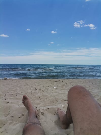 The image depicts a first-person perspective of a person lying on a sandy beach, with their legs visible in the foreground. The sand is light brown and appears undisturbed, with some scattered debris such as small pieces of wood and a shell. The ocean stretches out in the middle ground, with moderate waves and a clear horizon. The sky is a bright blue with scattered white clouds. The location is the beach in Osenovo, Bulgaria.