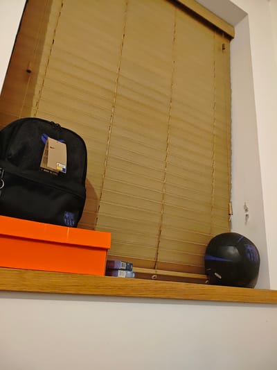 A still life is composed of a black backpack with a tan tag on top of an orange Nike shoebox; two small, blue boxes; and a black soccer ball, all sitting on a wooden window sill. The background consists of a closed, light brown, horizontal slat blind. The location is Swindon, United Kingdom. No people are present.