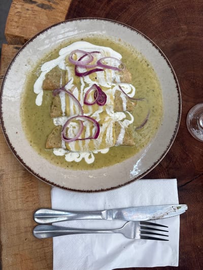 A plate of four green enchiladas, topped with white cream and slices of red onion, rests on a light brown wooden surface with prominent grain. The enchiladas are submerged in a light green sauce. To the lower left of the plate, a silver-colored knife and fork are positioned on a folded white paper napkin. A portion of a clear glass stemware item is visible on the upper right side of the wooden surface. The scene is located in Santiago Cuautlalpan, Mexico. No individuals are present in the image.