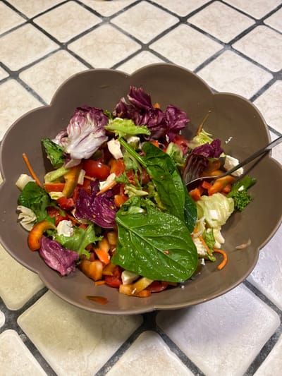 An overhead photograph captures a brown, scalloped-rimmed bowl filled with a mixed salad, positioned on a light-toned tiled surface with dark grey grout forming a diamond pattern. The salad contains various types of lettuce, including light green leaves and prominent red-purple radicchio. Other visible components include diced red tomatoes, chopped red and orange bell peppers, julienned carrots, and small, irregular white cubes consistent with cheese. A single large, dark green, glossy leaf, likely basil, rests prominently on the salad's surface. A silver-colored metal fork is partially visible within the salad on the right side of the bowl. The scene is located in Cava de' Tirreni, Italy.