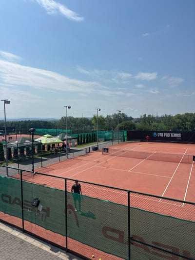 This image captures an outdoor tennis complex located in Giebułtów, Poland, under bright, clear daylight with a mostly blue sky and scattered high cirrus clouds. The perspective is elevated, looking down upon the facilities.
In the immediate foreground, a black metal fence topped with a green mesh banner, displaying "HEAD" branding, partially obstructs the view. Two yellow tennis balls rest on the paved ground directly in front of this fence. A male individual, wearing a dark top and light-colored trousers, is partially visible above the green banner, walking from left to right along the paved perimeter, casting a distinct shadow. A small, black wheeled device is attached to the green mesh banner.
The mid-ground features multiple reddish-brown clay tennis courts, defined by white lines and equipped with white nets. The nearest court is unoccupied. On the court beyond it, a male individual dressed in a red shirt stands near the far baseline, facing away from the viewer, appearing to be engaged in tennis activity. A large black banner at the back of this court displays "UTR PRO TENNIS TOUR" in white text. Tall light poles with square floodlights are positioned around the court areas.
To the left of the main courts, several pop-up tents are erected on a paved area: one green tent with "TIMBARK" branding, and two darker tents. Several indistinct figures are visible beneath these tents. Further into the background, additional tennis courts are visible amidst green fencing and surrounding trees. The horizon reveals distant rolling hills and dense green foliage, suggesting a rural or semi-rural environment beyond the complex.