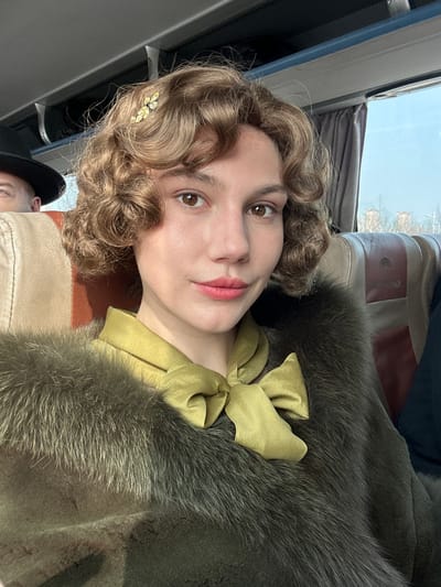 A female individual, estimated in her 20s or 30s, is centered in a close-up selfie taken inside a King Long brand passenger bus, presumed to be in Huangdao District, China. She possesses fair skin, brown eyes, and light brown hair styled in voluminous curls with a gold-toned, leaf-shaped hair clip adorned with clear stones on the right side of her head. Her lips are coated with pink lipstick. She wears a golden-olive green satin blouse featuring a large bow tied at the neck, overlaid by a dark olive-green fur coat or stole. Behind her, the upper portion of a second individual, wearing a dark brimmed hat, is partially visible. The bus interior features beige and brown upholstered seats, an overhead compartment, and a window with a drawn curtain. Through the window, a clear blue sky, distant low-rise buildings, and sparse trees are visible, indicating an exterior environment.