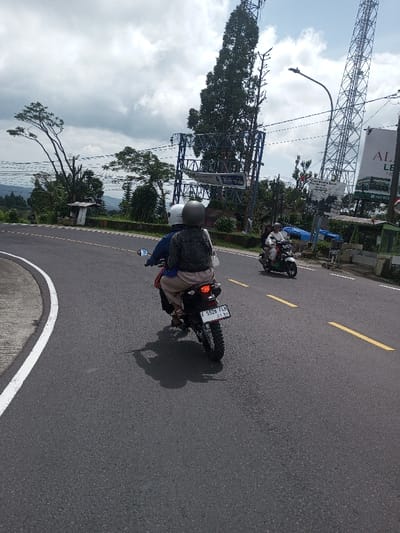 The scene is located in the city of Gubbisamamidi, India, depicting an outdoor road on a partially cloudy day. A two-lane asphalt road, marked with a solid white line on the left curve and broken yellow lines in the center, dominates the foreground and midground. Two motorcycles are traveling away from the viewer.
In the immediate foreground, a black dirt-bike style motorcycle is seen from the rear, navigating a gentle left curve. The motorcycle carries two individuals. The pillion rider, situated behind the driver, wears a dark gray helmet, a dark gray denim-like jacket, and light beige pants, with a white bag slung over their left shoulder. The driver, partially obscured, wears a white helmet and a blue long-sleeved shirt. The motorcycle's license plate reads "F 5526 FLH 07.23" and its taillight is illuminated. A shadow of the motorcycle and riders is cast on the road.
Further down the road on the right, a light-colored scooter or smaller motorcycle is visible, also traveling away from the viewer. This motorcycle carries at least two, possibly three, individuals, both wearing helmets. The driver is in a white top, and the passenger in a dark top.
The right side of the road features various structures and vegetation. Two large, metal billboard frames are prominent, one partially obscured by a tall, slender coniferous tree. A blue banner with "3GB 99rb" is visible on one billboard. Another sign reads "ALAM..." and part of a phone number "+62 823...". A tall, lattice-style telecommunication tower stands behind these signs. A streetlight on a pole is also present. Several small, makeshift stalls with blue tarpaulins and metal roofs are located along the roadside.
To the left of the road, beyond a dark green barrier, dense vegetation and trees are visible. In the distant background, a mountainous or hilly landscape extends under a sky with significant cloud cover and patches of blue.