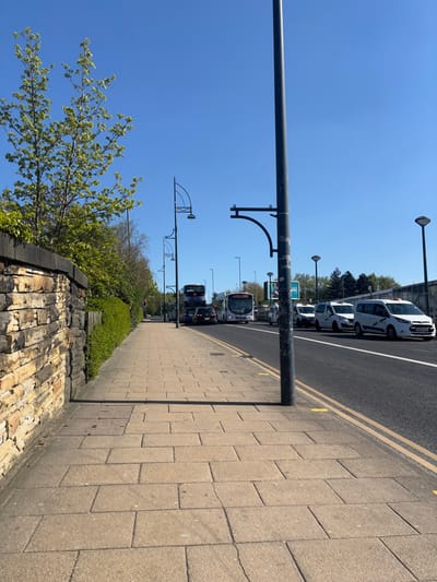The image shows a sidewalk and street in Bradford, UK, at latitude 53.791 and longitude -1.751. The sidewalk is paved with gray concrete blocks and runs parallel to the street. A stone wall is on the left side of the sidewalk. There are trees and bushes along the sidewalk. On the street, there are several vehicles, including two double-decker buses, a white van, a white sedan, and a taxi. The street is paved with asphalt and has a double yellow line down the center. There are several streetlights along the sidewalk and street. The sky is blue and cloudless. The time of day is likely afternoon.