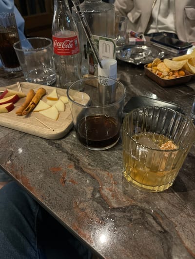 The image captures a table setting in an indoor environment, likely a restaurant or bar, with a variety of items. A glass of amber liquid sits near another glass holding a dark liquid. A platter contains cheese slices, breadsticks, and apple slices. There's an empty Coca-Cola bottle, an open container of chips, and some crumpled napkins. The table has a textured, grey-brown surface. The image was taken in Xanthi, Greece.