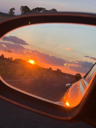 The image shows a sunset viewed in the reflection of a car's side mirror. The sky is painted with vibrant orange, pink, and purple hues, with scattered clouds. The sun, a bright orange orb, is low on the horizon, casting a warm glow over the landscape. The landscape appears to be rural, with rolling hills and trees visible in the distance. A road is reflected in the mirror, with a few cars driving on it. The car's body is also visible in the foreground, with its side mirror in focus. The image evokes a sense of peace and tranquility. The location is described as Дудзіцкі сельскі Савет, Belarus.