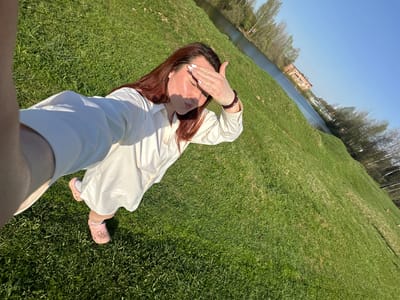A woman lies on green grass near a body of water, potentially a canal or river. She is wearing a long white dress or shirt, pink Crocs, and a bracelet. She has reddish-brown hair and her hand is raised to shield her eyes from the sun. In the distance, a brick building is visible across the water. The location is Черёмушки, Russia.