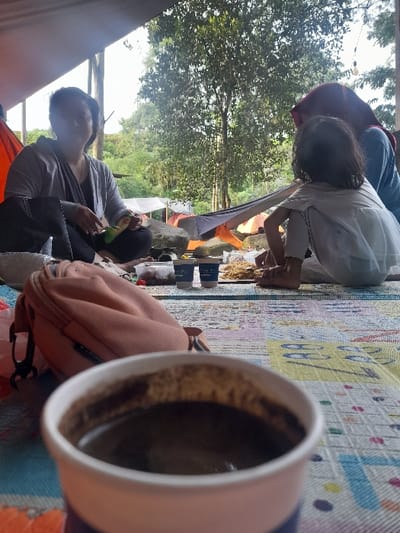 A campsite scene features a woman in a gray shirt and black top eating inside a tent. Near her is a brown backpack. A child with dark hair wearing a white shirt and purple hoodie sits on a colorful mat beside a woman. In the foreground, an out-of-focus mug of coffee sits on the mat. The background shows lush green trees. The photo appears to be taken from within the tent, capturing a casual outdoor moment.