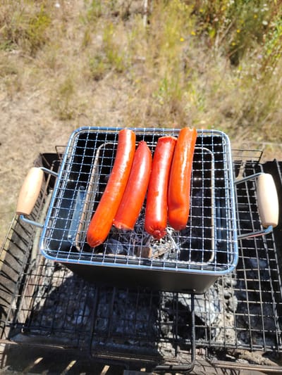 Four bright red sausages are arranged on a chrome grilling rack over hot coals. The rack sits inside a black, rectangular charcoal grill. The grill has two wooden handles on either side. The background shows dry, yellowed grass and some out-of-focus green foliage under a clear, sunny sky. This scene is located in the city of Staiky, Ukraine.