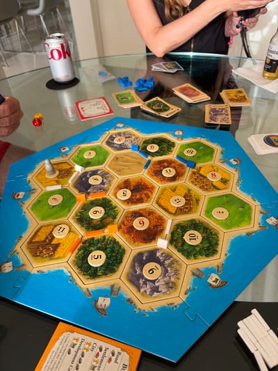 The image depicts a game of Settlers of Catan in progress, likely at a residence. Visible components include the game board with hexagonal resource tiles, number tokens, roads, settlements, and at least one city. Also visible are development cards and resource cards. A red Coke can, Corona beer bottle, and dice are visible. People can be seen at either side of the board.