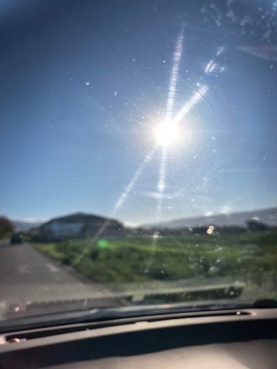 The image appears to be taken from inside a vehicle looking out through the windshield. The sun is bright and high in the sky, obscuring details due to the glare. The windshield is dirty, with smudges and streaks. In the background, there is a building or structure, a green field or grassy area, and what appears to be a road. The location is likely Krainitsi, Bulgaria.