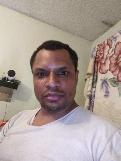 A head-and-shoulders, selfie-style photograph depicts an adult male indoors. The subject, an individual with dark skin, short dark hair, and a short beard, is centered, looking directly into the camera with a neutral expression. He is wearing a light-colored, likely off-white or light grey, short-sleeved t-shirt with a wide neckline. The immediate background on the left features a light-colored wall, possibly pale green, with a wall-mounted shelf or bracket holding a small white cylindrical object and associated wiring. A brown, cardboard-like item is visible below this shelf. To the right background, a floral-patterned fabric, displaying large red/pink roses and green foliage on a light base, is draped or hung against the wall. The ceiling is textured, appearing light in color. The scene is consistent with a residential interior within the city of Shoreline, United States of America.