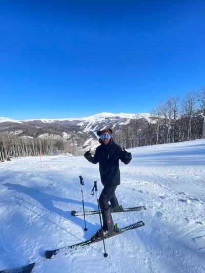 The image depicts a male individual standing on a snow-covered ski slope under a clear blue sky, presumably in the vicinity of Mušovića Rijeka, Montenegro. The individual is centrally positioned, facing the camera, and performing a "thumbs up" gesture with both hands. He is dressed in winter ski attire, including a black hooded jacket, black ski pants, ski goggles, and a brown head covering with prominent, rounded, fur-like ears. He is wearing skis with green bindings and black ski boots, with ski poles partially visible. The foreground shows groomed snow with visible ski tracks. In the midground, a red marker pole is present on the left side of the slope. The background features extensive snow-covered mountain ranges with forested slopes, primarily composed of bare deciduous trees, under a bright, cloudless sky. Long shadows are cast on the snow, indicating direct sunlight.