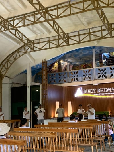 The image shows an indoor assembly area with a large, light-colored wooden sign with the words "BUKIT RHEMA House Of Prayer For All Nations" in white and gold lettering. There are several men standing at the front of the room, some holding microphones, addressing a group of people seated in rows of wooden benches. Some people are also standing near the back of the room. The ceiling features a crisscrossed pattern of exposed brown beams and a painted sky, reminiscent of a traditional Indonesian architecture. The room is located in the Bukit Rhema prayer house in Kecamatan Borobudur, Indonesia, at coordinates -7.606, 110.18.