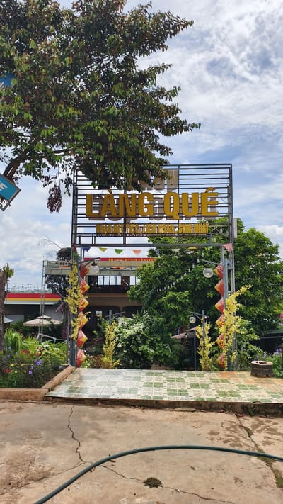 This image shows a sign for "LÀNG QUÊ" which translates to "Countryside" in Vietnamese, suggesting a rural-themed establishment. The sign is prominent, with large yellow letters on a dark metal frame, set against a bright, partly cloudy sky. Below the main sign, smaller text is visible, including "NHÀ HÀNG TIỆC SINH NHẬT" which means "Birthday Party Restaurant" and "ẨM THỰC CAFE LÀNG QUÊ" which translates to "Countryside Cuisine Cafe".

The setting appears to be an outdoor entrance to a restaurant or cafe. There is a tiled pathway leading towards the establishment, with decorative elements like strings of lights and hanging lanterns, some adorned with yellow flowers. Lush green trees and flowering bushes surround the entrance, creating a vibrant and inviting atmosphere. In the background, a multi-story building with awnings and windows is visible, along with umbrellas for outdoor seating. The overall impression is a pleasant daytime scene, likely in the late morning or afternoon given the bright lighting and clear sky with scattered clouds. The location context of Ia Hru, Vietnam, suggests this is a local establishment in a rural or semi-rural area of the country. No people are visible in the image.