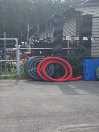 In the town of Deulino-3, Russia, an outdoor storage area showcases construction materials. In the foreground, two coiled corrugated pipes, one dark gray and the other bright red, are placed on asphalt. Behind them, a wire mesh fence encloses various items including stacked pipes and metal shelving units. Beyond the fence, a brick building with a dark roof stands, with large, ribbed black cylindrical structures positioned against its side. Several small white signs are attached to a horizontal bar in front of these structures. To the right of the building, a bright blue plastic container is visible, along with a section of a green manhole cover on the ground. Lush green foliage grows around the building and extends behind the fence. The scene appears to be bathed in natural daylight, with some shadows cast on the asphalt.
