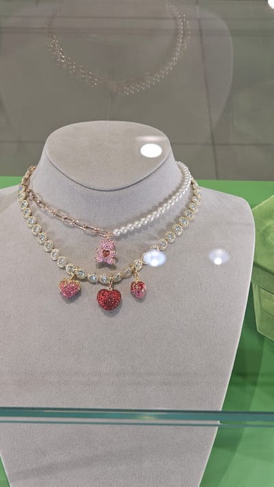 Two necklaces are displayed on a gray mannequin bust in a display case. The upper necklace consists of a strand of small white pearls. The lower necklace is a chain with clear cubic zirconia stones and a rose gold chain link section. Both necklaces feature dangling charms. The lower necklace has three charms: a pink heart-shaped strawberry, a larger red heart, and a smaller pink strawberry. A pink teddy bear charm is suspended from the rose gold chain link section of the lower necklace. The display case has a glass front, reflecting a blurred image of another necklace above the mannequin. The background includes a green surface. This scene is located in a jewelry store in Zaragoza, Spain.