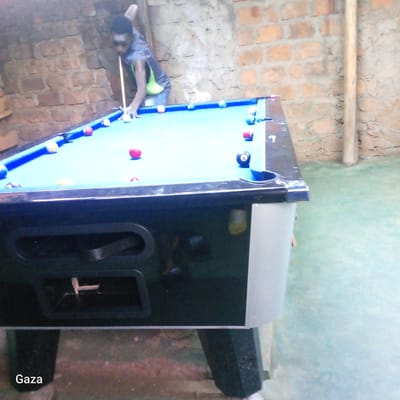 An individual is depicted playing pool indoors in Kamera ward, Kenya. The person, appearing male and of African descent, wears a grey short-sleeved shirt with a bright underlayer and blue jeans, positioned with their torso bent over a pool table. They are holding a light-colored cue stick with their right hand, while their left hand forms a bridge on the table's light blue felt surface. A white cue ball is visible in front of the cue stick, with numerous colored and striped pool balls scattered across the table, including several solid red balls and a black 8-ball. The pool table features dark blue rails and a black and grey body. The background consists of an unfinished brick wall, characterized by reddish-brown bricks and visible mortar. A light-colored vertical post stands against the wall on the right. The floor is a light green or faded blue concrete surface. The word "Gaza" is visible as white text in the bottom left corner of the image.