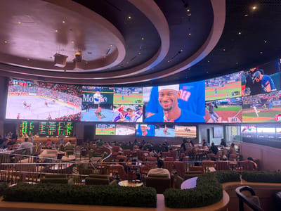 The image depicts a sports betting lounge, likely located in Las Vegas, Nevada, USA, due to the presence of sports screens and betting odds displays. The lounge features a multi-screen display system showing various sporting events, including hockey, baseball, horse racing, and mixed martial arts. Patrons are seated at tables and in lounge chairs, watching the screens, and presumably placing bets. The environment appears upscale with tiered seating, decorative greenery, and modern lighting. The overhead lighting fixtures suggest a high-end interior design. People are generally relaxing, watching sports, and interacting in small groups. The general environment is busy and active, indicating a popular establishment.