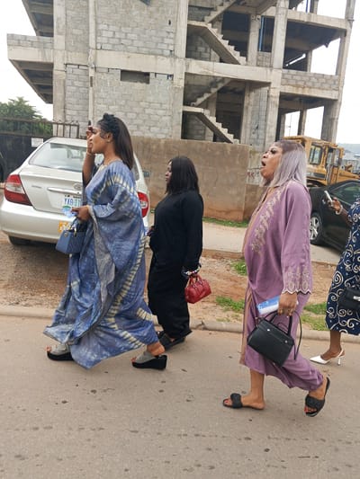 The image shows a street scene in Guzape, Nigeria, with a partially constructed building in the background. Three women are walking on a paved road. The woman in the foreground on the left is wearing a blue and white tie-dye patterned dress and grey platform sandals. She is carrying a blue handbag. The woman in the middle is wearing a black outfit and red flat shoes, and she is carrying a red handbag. The woman on the right is wearing a purple patterned dress and black slip-on sandals, and she is carrying a black handbag. A silver Toyota car is parked on the left side of the road. A yellow construction vehicle is visible in the background.