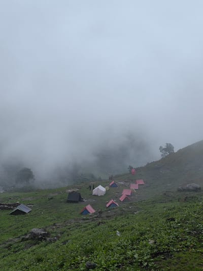 The image depicts a campsite in Dhundi, India, nestled within a foggy mountain landscape. Numerous tents, in various colors including red, blue, white, and black, are scattered across a grassy slope. Some tents are clustered together, while others are more isolated. Two individuals are visible near a white tent on the upper right side of the frame, appearing to be engaged in some activity. The environment is characterized by lush green vegetation, rocks, and a thick, pervasive fog that obscures the background and creates a soft, diffused light. The overall impression is one of a remote and serene mountain settlement.