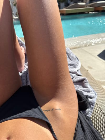 A tanned individual lies by a pool. They are wearing a black swimsuit. The image focuses on their upper thigh, where stretch marks are visible. A small tattoo of text in a vertical orientation is present. A gray towel rests nearby. The pool is visible in the background.