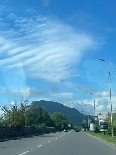 The image depicts a view of a road leading towards a mountain range, with a clear blue sky dotted with white clouds. On the right side of the road, there are streetlights and billboards. One billboard displays text in Russian and images of bottles. A traffic sign indicating a speed limit of 40 is also visible. On the left side of the road, there is a decorative fence and trees, with a bare tree in the foreground. Several cars are driving on the road. The overall scene suggests an urban or suburban environment on the outskirts of Mineralnye Vody, Russia, with the mountains serving as a prominent natural backdrop.