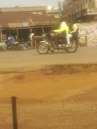 In Nabatum, Nigeria, a motorcycle carrying two individuals travels down a dirt road in front of a row of buildings with corrugated metal roofs. The rider wears a neon yellow jacket and dark pants, while the passenger is in a light-colored shirt and white pants. Another motorcycle with a rider in a blue jacket is parked to the left, and another motorcycle's front wheel is visible on the right side of the frame. The ground is a mixture of dirt and gravel, with a raised, eroded embankment on the right. The buildings appear to be shops or small businesses, with open doorways and some items visible inside. The overall scene depicts a busy street environment with everyday activity.