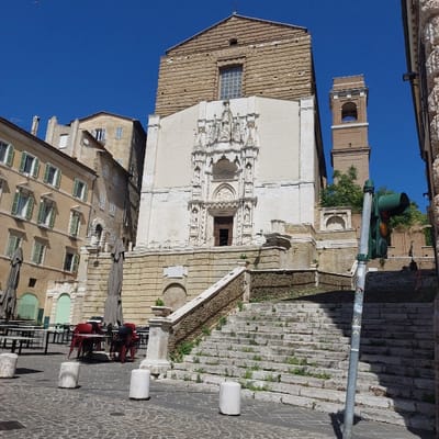 This image depicts the Church of San Francesco alle Scale, located in Piazza San Francesco alle Scale in the city of Ancona, Italy, under a clear blue sky.

**Scene Description:**
The central subject is the Church of San Francesco alle Scale, presenting a two-tiered facade. The lower portion is a white, intricately carved Gothic portal with statues and a central arched entrance, positioned above a substantial stone base. The upper portion of the church is constructed from large, rough-hewn, light-brown stone blocks, featuring a single rectangular window and a plain gable roof topped with a cross. To the right of the main church structure, a tall, slender brick bell tower with arched belfry openings stands.

A wide, multi-tiered stone staircase, exhibiting some vegetation growth between its steps, leads from the cobblestone square up to the church entrance. The staircase features stone balustrades on its left side (viewer's perspective) and is partially shaded on the right.

In the immediate foreground, a cobblestone square is visible. Three white, cylindrical concrete bollards are spaced across this area. To the left of the staircase, an outdoor seating area with several tables (some square, some round) and stacked red chairs is present, alongside multiple large, gray, folded outdoor umbrellas. To the right, a dark green vertical traffic light pole with an illuminated green signal is positioned, partially obscuring the lower steps of the staircase and casting a shadow.

In the background on the left, several multi-story historic buildings with light-colored facades and numerous windows with green wooden shutters ascend a gentle slope. On the far right, behind the bell tower, a terraced hillside or retaining wall with sparse vegetation is visible. No individuals are present in the scene, and no actions or interactions are depicted. The environment suggests bright daylight.