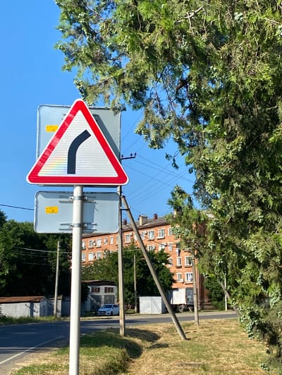 A traffic sign indicating a sharp right turn is positioned on a white pole. The sign is a red-bordered triangle with a black curved arrow pointing right. Below this sign, another rectangular sign with a yellow rectangle is attached to the same pole. The background features a clear blue sky, dense green foliage from a tree in the foreground on the right, and a building with multiple windows, appearing to be an apartment complex, in the mid-ground. Power lines are visible in the sky. A paved road runs horizontally across the lower part of the image, with dry grass and some vegetation on the side. A white car is parked on the right side of the road in the mid-ground. The overall scene depicts a street in a city or town, possibly in a suburban area, under daylight conditions.