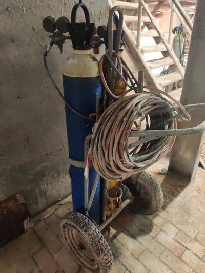 The image shows a mobile gas cylinder cart, likely used for welding or cutting, situated in an industrial or workshop setting. The cart is blue and yellow and features two large wheels at the back. A thick, coiled hose, appearing to be a welding hose, is draped over the front of the cart. Attached to the cylinder are regulators and hoses, suggesting it holds a pressurized gas. In the background, a rusty metal staircase is visible, along with concrete walls and a brick floor, all contributing to a utilitarian and somewhat weathered environment. There are no people in the image, and the lighting suggests an indoor setting, possibly during daylight hours though the exact time of day is not discernible. No visible text is present on the main subjects of the image. The overall impression is one of a functional, albeit not pristine, workspace. The presence of industrial equipment and the general state of the surroundings imply a location in Poland, likely a workshop or a construction site.