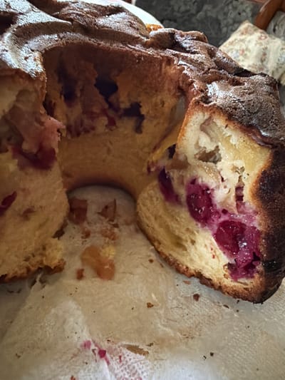A bundt cake, partially eaten and with a slice removed, sits on a white cloth. The cake has a golden-brown crust and a soft, light-colored interior. Visible within the cake are chunks of what appear to be dark red berries, likely cherries or cranberries, and possibly some lighter fruit pieces. Crumbs and a faint pink stain are present on the cloth, suggesting the cake has been served. In the background, out of focus, is a floral patterned fabric. The setting is indoors, likely a domestic environment. There is no information provided about the city of Манихино Снт, Russia in relation to this image.