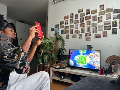 A man wearing a black silk robe with embroidered dragons, white pants, and a black satin durag sits in a room, holding a red smartphone up to take a picture. He has dark skin, a mustache, and a ring on his left hand. The room appears to be a living space. Behind him is a white wall adorned with numerous framed prints of various artworks. A leafy green potted plant sits to the left of a white TV stand, atop which rests a flat-screen TV displaying a video game with cartoonish characters. A fan and a small potted cactus sit to the right of the TV. The TV stand is also holding various objects including a Nintendo Switch, papers and books, and a remote control. The floor is made of light brown wood. There is a glimpse of white kitchen cabinets in the background on the left. There is no clear evidence to confirm this photo was taken in Pantin, France.