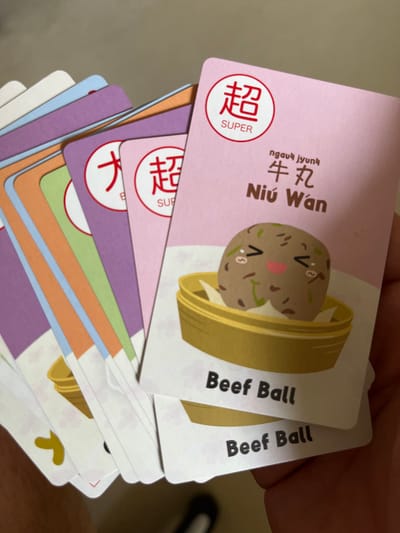 A hand holds a fanned-out stack of colorful cards, likely flashcards or game cards. The top card, predominantly light pink, features a charming cartoon illustration of two beef balls (牛丸 / Niú Wán) resting in a bamboo steamer basket. One of the beef balls has a cute, smiling face with rosy cheeks, conveying a cheerful emotion. In the upper-left corner of this card, a red circle displays the Chinese character "超" (chāo) above the English word "SUPER". Alongside the illustration, the card presents the text "ngauk jyunk", "牛丸", and "Niú Wán". Vertically arranged on the right side of the card, the English translation "Beef Ball" is clearly visible, repeated for the card directly underneath.

Below the main pink card, a variety of other cards are fanned out, showcasing different colors such as purple, green, orange, and light blue. Another red circle with "超 SUPER" is partially visible on one of the underlying cards. The visible text indicates a bilingual set, presenting information in both Chinese and English, suggesting an educational purpose or a game. The activity taking place is simply holding and displaying these cards. The scene appears to be indoors, with no specific time of day or weather discernible. No people are visible, only the hand holding the cards. This image was captured in Middle Park, Australia.