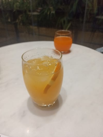 The scene is located in San Marzano sul Sarno, Italy. A white, circular tabletop occupies the lower two-thirds of the image. Positioned in the foreground, slightly left of center, is a clear, stemless glass containing a light amber liquid. Multiple ice cubes and a single orange or citrus wedge are visible within the liquid, which exhibits surface effervescence and small bubbles adhering to the interior glass wall. Further back and to the right, a second, smaller, clear, stemless glass, out of focus, holds a darker orange liquid. The background beyond the table is dark and blurred, revealing indistinct green foliage, indicative of an evening or low-light outdoor or semi-outdoor environment. No individuals or active interactions are depicted; the scene presents a static arrangement of beverages.
