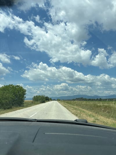 The image depicts a point-of-view shot from inside a vehicle, looking out through the windshield at a rural road in Novo Zhelezare, Bulgaria. The road, paved and marked with a white line, extends into the distance, flanked by dry, golden grass and scattered green trees. In the background, a range of low, blue-tinged mountains is visible under a vast sky filled with dramatic, puffy white clouds. The foreground shows the dark dashboard of the vehicle, obscuring the lower portion of the scene. There are no people or animals visible in the image. The environment suggests a clear, sunny day in a rural, possibly agricultural, area.
